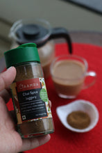 Hand holding a spice jar labeled 'Chai masala' on a red surface with containers in the background.