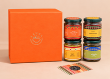 An orange gift box with a white logo next to four glass jars of chutneys and a small card, photographed on a warm beige background. The jars have colorful labels and dark lids.