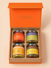 An orange gift box containing four jars of spices arranged in a 2x2 grid. The jars have colorful labels and are set against a solid light beige background.
